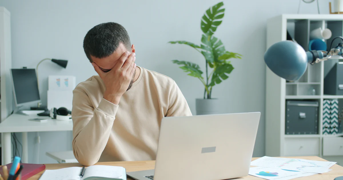 Homem adulto com a mão na testa diante do notebook, com dificuldade de concentração no trabalho