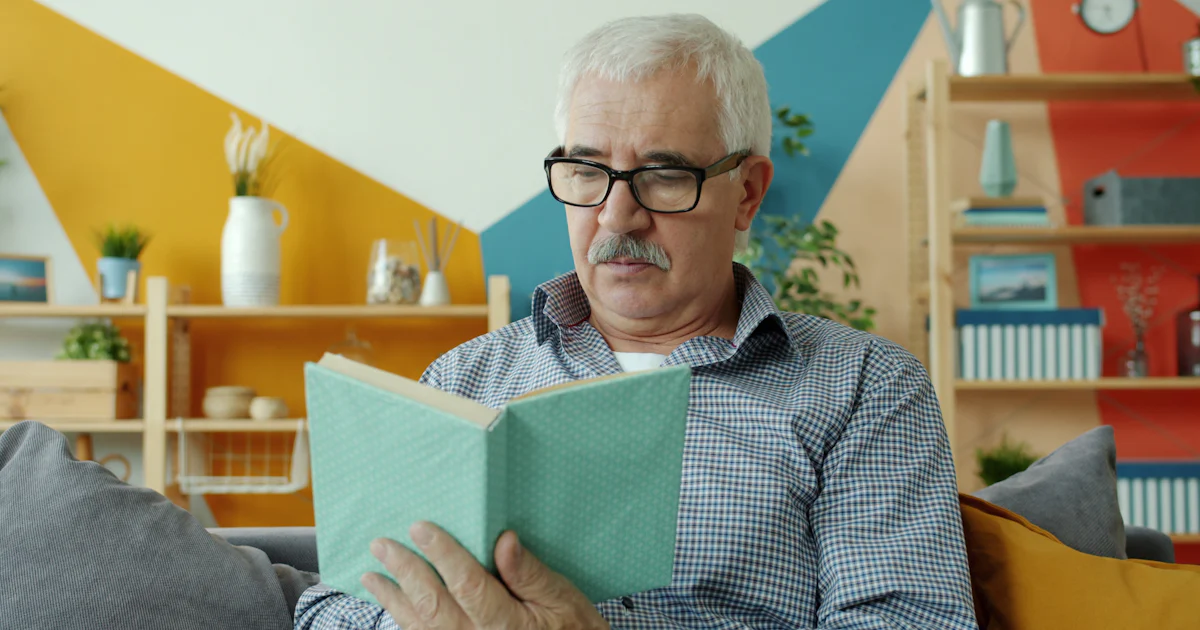 Homem idoso de óculos lendo um livro no sofá de casa