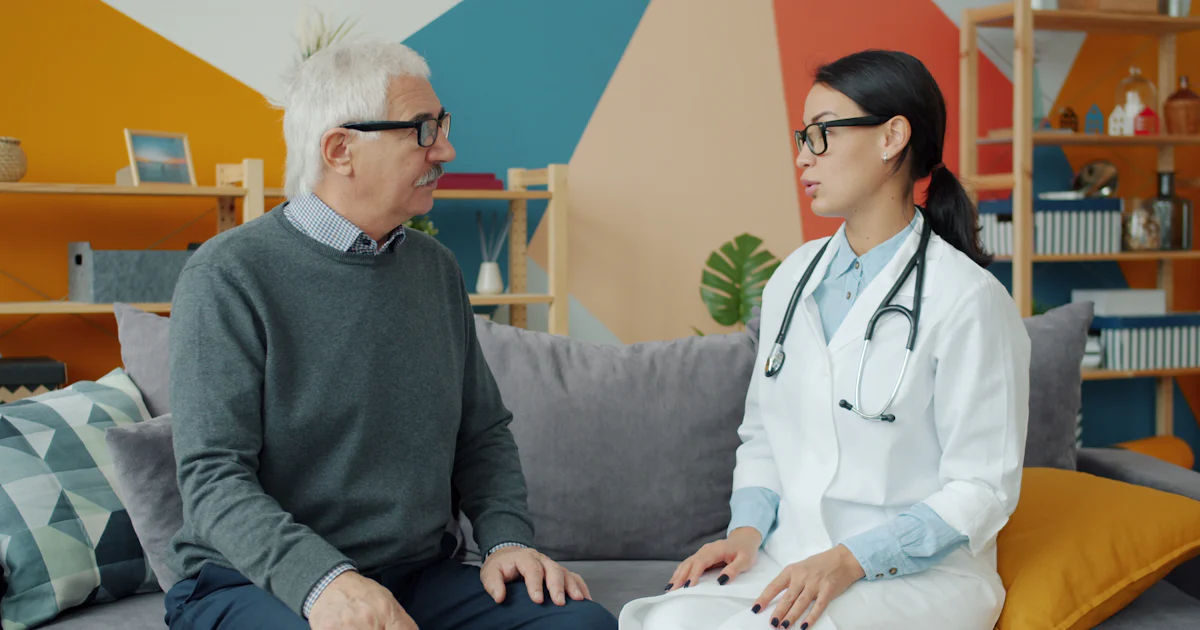 Médica conversando com um homem idoso, os dois sentados no sofá