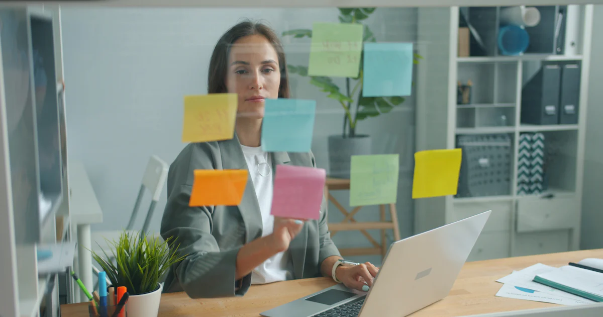 Mulher organizando tarefas em notas adesivas coloridas enquanto trabalha no notebook